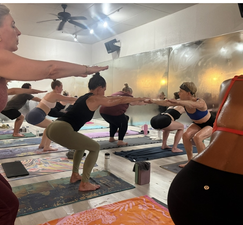 A packed Hot Yoga class holding chair pose, mirrors fogged with steam